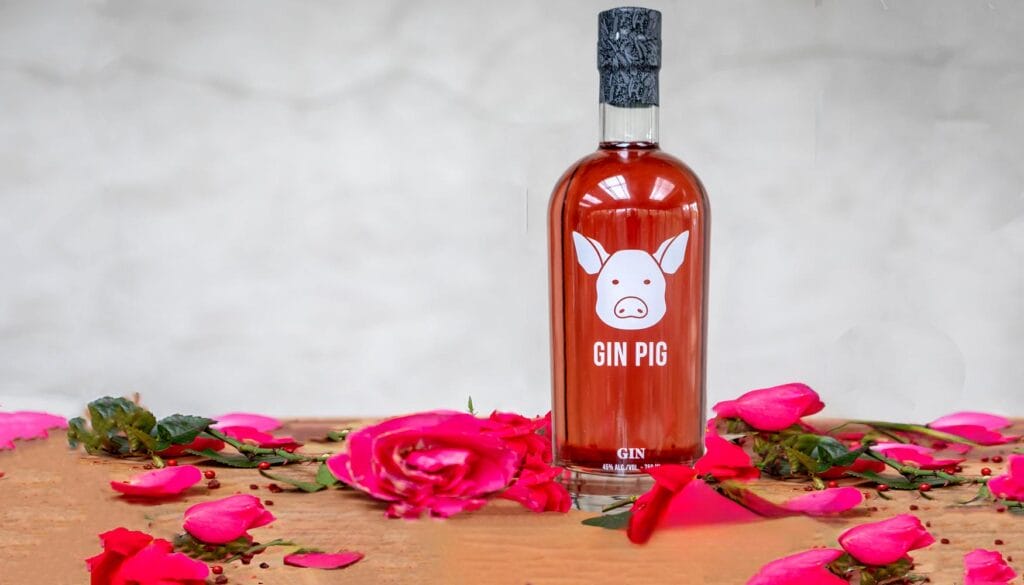 A hand pouring a bottle of CW gin into a glass filled with ice and garnished with a lime wheel, surrounded by pink rose petals and peppercorns.